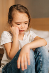 Close-up face of distraught beautiful little girl with wet eyes from tears sits on bed in bedroom...