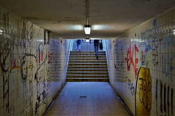 Some people walk up the steps of a dark and dingy subway tunnel beneath a railway line, with the walls covered in spray paint and graffiti