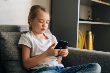 Close-up of little girl talking on video call by mobile phone in living room sitting on cozy sofa. Adorable child looking fun video on cellphone sitting on couch. Concept of leisure activity at home