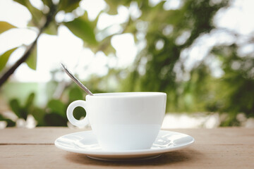 A white hot coffee cup with spoon in a cup and saucer are placed on a wooden plate and on the landscape nature background.