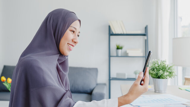 Young Asia muslim businesswoman using smart phone talk to friend by videochat brainstorm online meeting while remotely work from home at living room. Social distancing, quarantine for corona virus.