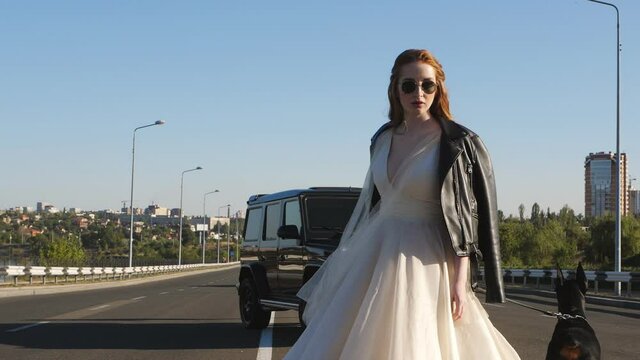 The bride with a Doberman in a leather jacket and glasses turns to the camera on the roadway.