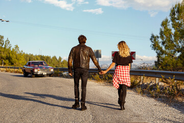 Back view of couple wearing rock style casual clothing walking towards their vintage red muscle car parked on the side of the winding road with beautiful mountain view. Copy space, background.