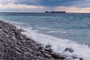 Fototapeta na wymiar The cargo ship sails on the sea horizon. Stone coast and sea waves in the foreground