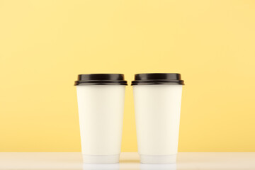 Two white tall disposable coffee or tea cups on white table against light yellow background with copy space. Hot drinks minimalistic concept