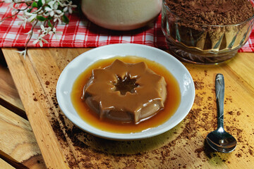 Chocolate pudding on porcelain plate. Milk jar and glass jar with chocolate in po. Wooden base with checkerboard fabric. Black background.