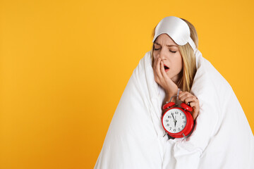 Young woman in sleeping mask wrapped with blanket holding alarm clock on yellow background. Space for text