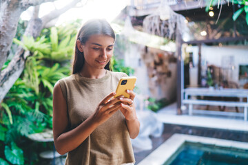 Positive young ethnic woman using smartphone near resort pool