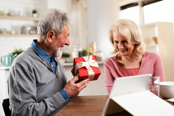 Senior couple having video call. Happy husband giving his wife a gift.