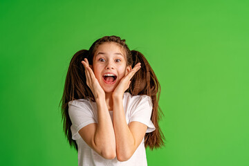 cheerful girl about 10 years old in white T-shirt holding her hands near her face and happy smile on green background. happy child enjoys success and victory. Discounts and sales. Advertising concept