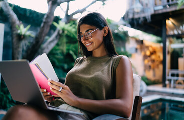 Gorgeous ethnic lady using netbook and reading diary while working remotely on terrace