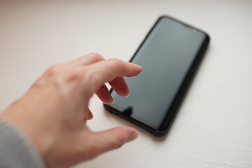 Female hand reaching for black smartphone on white table.