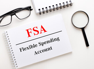 FSA Flexible Spending Account is written in a white notebook on a light background near the notebook, black-framed glasses and a magnifying glass.