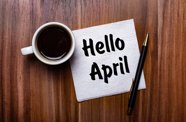On a wooden table next to a white cup of coffee and a pen is a white paper napkin with the words HELLO APRIL