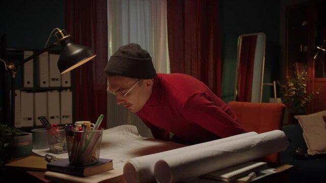 Young designer is sitting down and begining to draw plan with ruler and compass, making notes in project. Architecture student is learning at home in the evening. Creative space of architect. 