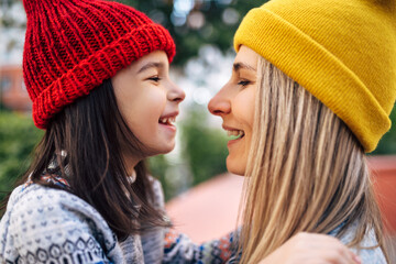 Side view portrait of cheerful little girl in red hat hugging her mom in yellow hat outside. Cute...