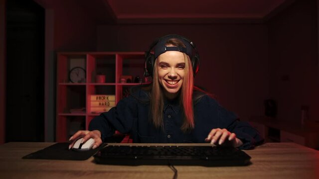 Portrait of a joyful lady gamer in casual clothes playing games at home on the computer at night and hiding from joy in a headset with a happy face. Positive female gamer playing.