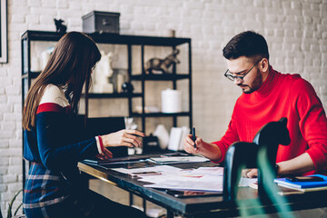 Naklejka na ściany i meble Male and female partners analyzing paperwork during brainstorming cooperation at table desktop, Caucasian couple 20s analyzing startup project success talking about report documents indoors