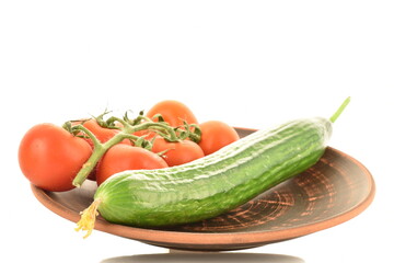 Several green cucumbers and red ripe, cocktail tomatoes on a twig on a clay plate, close-up, isolated on white.