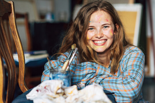 Candid portrait of a happy young woman with blots on her face smiling and looking directly to the camera after painting on canvas in her studio. Female artist have colorful paint strokes on her face.