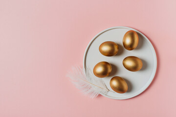 Golden eggs on white plate and feather on pink background, happy easter concept