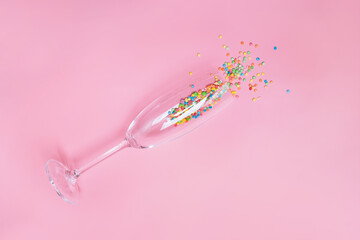 Colorful candy sprinkles in a champagne glass laying on pink pastel background.