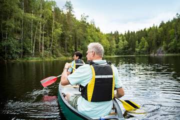 Fototapeta na wymiar Happy mature couple in life vests canoeing in forest lake. Sunny summer day. Tourists traveling in Finland, having adventure. 