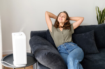 Fototapeta na wymiar Smiling and serene young woman rests on the comfortable couch under stream of fresh and cleaned air from humidifier in cozy living room, housewife leaned back with eyeclosed and daydreaming