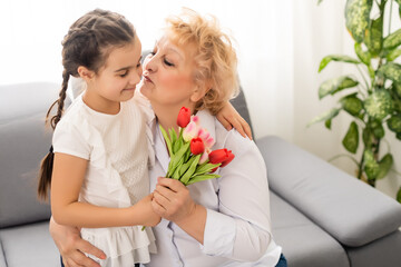 Fototapeta na wymiar Sincere cute little granddaughter strong cuddling and kissing in cheek her 60s grandmother gave her pretty spring flowers congratulates with birthday, International Womens day, close up concept image