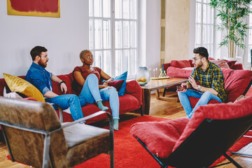 Diverse best friends dressed in casual wear communicating with each other resting at comfortable furniture in modern apartment, young hipsters talking indoors spending free time together at home