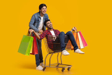 Two young men laughing with one of them sitting in a trolley holding carry bags.	