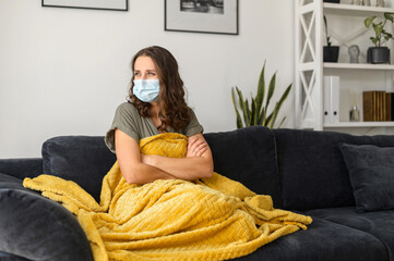 Young sick woman wearing protective face mask feels unwell at home, sits on the couch covered with...