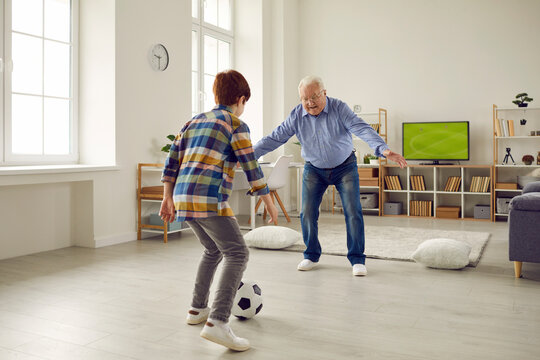 Happy Family Generation Bonding, Sport Game And Fun During Coronavirus Quarantine. Elderly Grandfather Playing Football With Preteen Grandson In Home Living Room Enjoy Spending Time Together