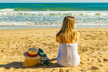 A girl is sitting on the sand with a covid mask and looking to the sea