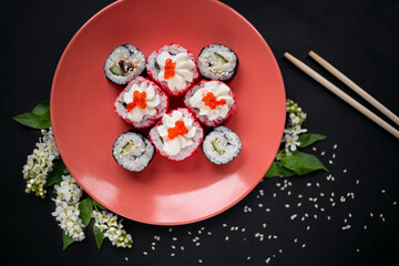 Top view of a sushi rolls set on a coral plate on a black background with flowers, sprinkled sesame and sushi sticks next to it.