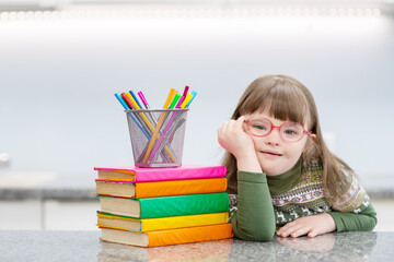 A girl with down syndrome at home leaned on a table heaped with books. Family education for children with disabilities