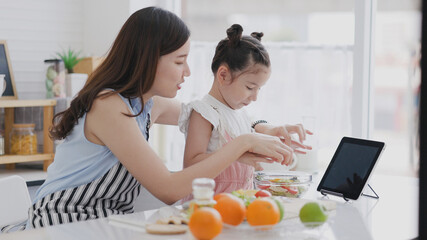 Family single mother and daughter enjoy and happy cooking in clean kitchen. Parents help children make fruit and vegetable salads from Video tutorial from tablet. Learn cook homemade from video call.