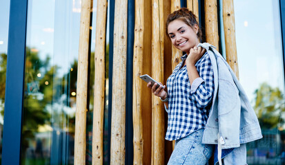 Half length portrait of cheerful millennial user dressed in stylish casual clothing smiling at camera during leisure for web networking, happy Caucasian blogger with smartphone posing outdoors