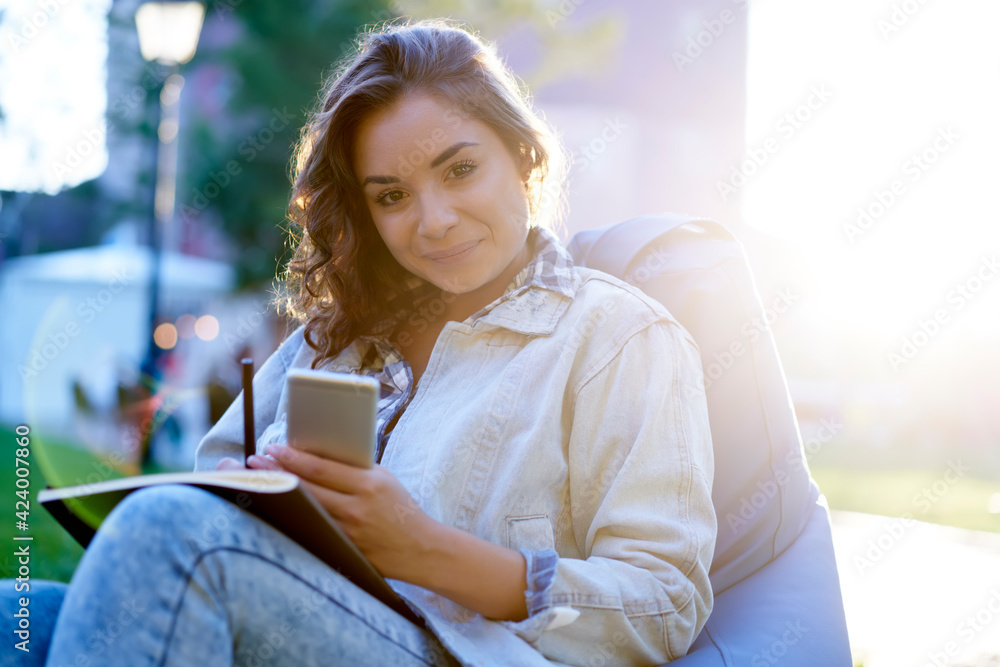 Poster backlit portrait of attractive female student with modern mobile phone and textbook for studying loo