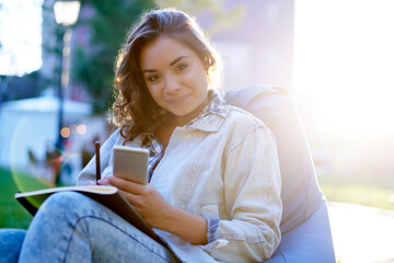 Backlit portrait of attractive female student with modern mobile phone and textbook for studying looking at camera during sunny day at nature, charming blogger with smartphone for networking posing