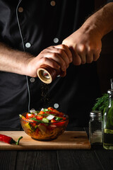A chef in a restaurant kitchen adds peppers to a fresh vegetable salad. Close-up of cook hands...