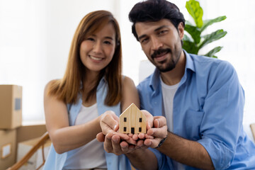 Smiling young family couple holding wooden home model in hands with cardboard boxes moving in a new home