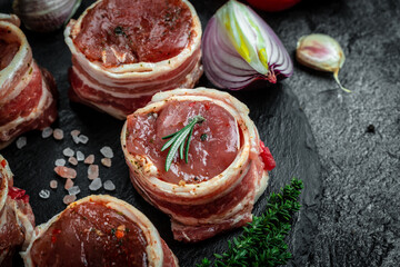 Raw pork medallion steaks wrapped in bacon served on a slate board on dark concrete background with spices salt and pepper, top view