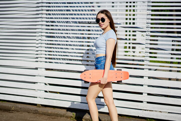 Obraz premium Portrait of millennial female skater in stylish sunglasses and casual apparel holding penny board for training skills, Caucasian teenage girl walking in city and posing during sunny day for rest