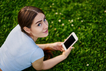Fototapeta premium Portrait of young Caucaisan woman holding digital smartphone in hands and looking at camera during free time, millennial hipster girl with modern cellphone device posing on green lawn grass in park