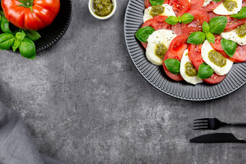 Traditional italian caprese salad with sliced tomatoes, mozzarella, basil, pesto sauce and spice on a dark background. Top view. Copy space.