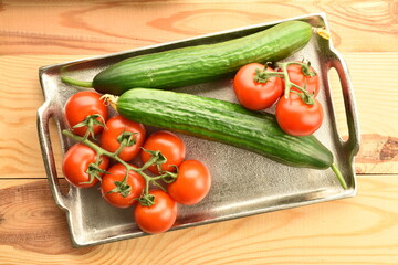 Several green cucumbers and red ripe, cocktail tomatoes on a branch with a metal tray, close-up, on a wooden table, top view.