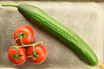 One green cucumber and three red ripe, cocktail tomatoes on a branch, close-up, on a metal tray.