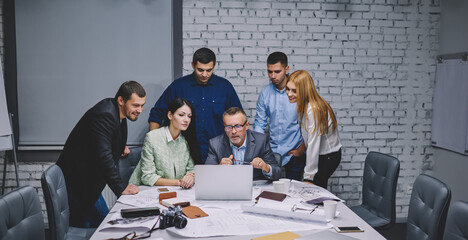 Experienced designers using modern laptop computer for searching online sketches and planning details of project, group of skilled architects collaborating at table desktop with netbook and blueprints