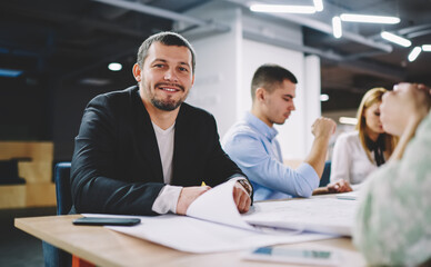 Portrait of happy Caucaisan businessman smiling at camera during collaborative meeting with professional colleagues enjoying positive brainstorming at table desktop, successful male employer